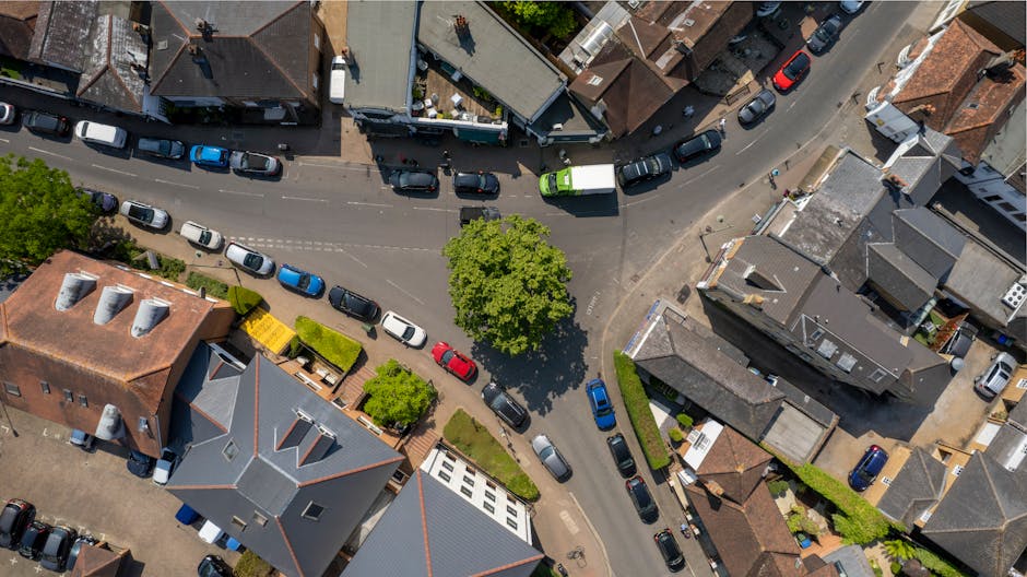 An aerial view of a residential street in TOLWORTH showing rows of parked cars lining both sides of the narrow road, with some vehicles close to the curb and others parked slightly further away. The street is surrounded by a mix of terraced and semi-detached houses with tiled roofs, some with chimneys, and small front gardens featuring shrubs and trees. A large, leafy tree stands prominently in the center of the image, casting a shadow over the parked cars and the pavement. The scene includes a vehicle partway through the loading process, likely a van operated by Man With a Van Tolworth, which is positioned on the pavement adjacent to the houses, with its rear doors open. Packing materials, such as cardboard boxes and bubble wrap, are visible on the pavement near the loading area, indicating a home relocation in progress. The environment is well-lit, suggesting daytime with clear weather, and the overall scene reflects the logistics involved in house removals along narrow streets, highlighting the importance of careful planning to avoid move-day delays, as addressed by [PAGE_TITLE].