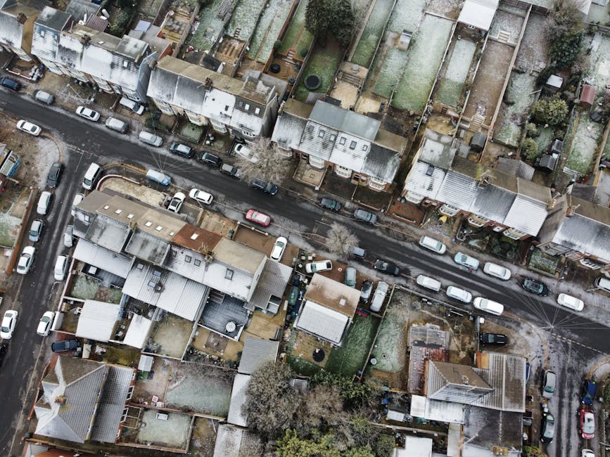An aerial view of a residential street in Tolworth with narrow roadways lined by closely spaced terraced and semi-detached houses, many with flat or gabled roofs, some showing signs of weathering. Parked cars, predominantly small and medium-sized, are aligned along both sides of the street, with several vehicles temporarily occupying the roadside during a home relocation process. The image captures the loading and unloading area where a man with a van service, such as Man With a Van Tolworth, is assisting with furniture transport and packing for a house move. The scene includes visible cardboard boxes, wrapped furniture pieces, and packing materials on the pavement and near the vehicles. The environment features small back gardens, some with trees and lawned areas, and features typical of suburban neighbourhoods in winter or early spring, with minimal foliage. Bright but diffuse daylight illuminates the scene, highlighting the logistical aspects of moving amid narrow street constraints typical of KT6 postal district, with the focus on safe loading, careful navigation, and efficient use of space during the home relocation process.