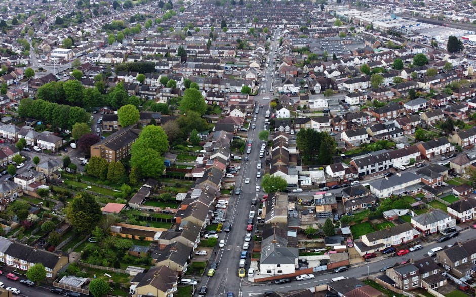 An aerial view of a residential street in TOLWORTH showing rows of parked cars lining both sides of the narrow road, with some vehicles close to the curb and others parked slightly further away. The street is surrounded by a mix of terraced and semi-detached houses with tiled roofs, some with chimneys, and small front gardens featuring shrubs and trees. A large, leafy tree stands prominently in the center of the image, casting a shadow over the parked cars and the pavement. The scene includes a vehicle partway through the loading process, likely a van operated by Man With a Van Tolworth, which is positioned on the pavement adjacent to the houses, with its rear doors open. Packing materials, such as cardboard boxes and bubble wrap, are visible on the pavement near the loading area, indicating a home relocation in progress. The environment is well-lit, suggesting daytime with clear weather, and the overall scene reflects the logistics involved in house removals along narrow streets, highlighting the importance of careful planning to avoid move-day delays, as addressed by [PAGE_TITLE].