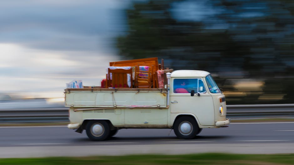 A vintage cream-colored van operated by Man With a Van Tolworth is shown during a home relocation process, traveling along a highway with a blurred background indicating motion. The van's cargo bed is loaded with various household furniture items, including a wooden dining table, matching chairs, cardboard boxes, and plastic bags filled with packing materials. The furniture is secured with straps or coverings to prevent movement during transit, and some items are wrapped in fabric or plastic for protection. The vehicle is positioned on the right side of the image, with the surrounding environment suggesting a rural or suburban setting illuminated by natural daylight. This scene exemplifies furniture transport and packing and moving logistics involved in house removals, highlighting the company's ability to handle bulky item removals efficiently during a home relocation in Tolworth.