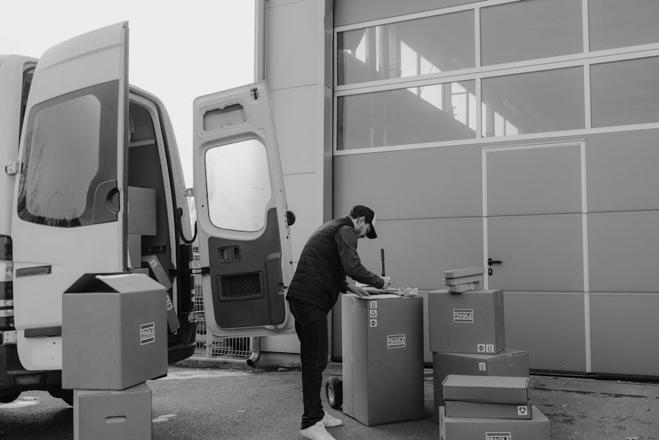 A man wearing a cap and a dark jacket is seen packing an area outside a modern industrial building with large glass windows and metal panels. He is standing next to a collection of cardboard boxes and