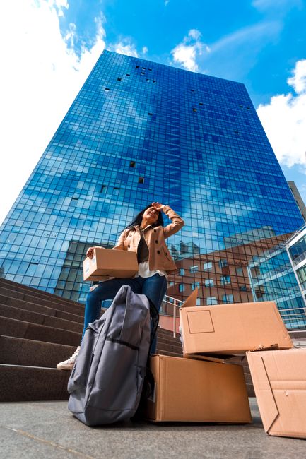 A man wearing a cap and a dark jacket is seen packing an area outside a modern industrial building with large glass windows and metal panels. He is standing next to a collection of cardboard boxes and