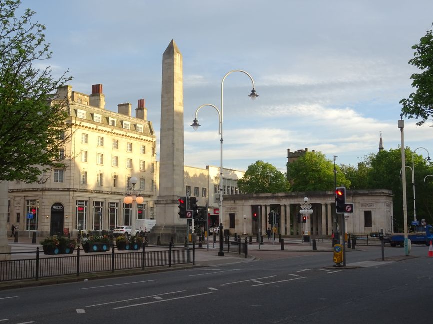 A view of a city street with a tall obelisk monument situated in the centre, surrounded by historic-looking white buildings with multiple chimneys on their rooftops. In the foreground, there is a road with traffic lights and pedestrian crossings, along with several street lamps and a few cars parked along the curb. To the right, a gated entrance with columns and a low stone wall is visible, and some people are walking nearby. The scene is set during daylight with a partly cloudy sky, and trees with green leaves line parts of the street, creating a balanced, urban environment. This setting is consistent with a typical city centre area suitable for home relocation or furniture transport activities managed by [COMPANY_NAME], providing a professional context for house removals and moving logistics.