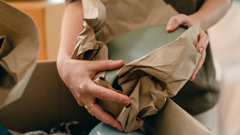 Close-up of a person's hands wrapping an antique item with a smooth, matte green surface in brown packing paper, preparing it for a home relocation. The individual is carefully securing the item inside a cardboard box using crumpled paper for padding, with additional packing materials visible nearby. The scene takes place indoors, with parts of furniture and soft lighting suggesting a residential setting. This detailed packing process, involving protective wrapping and precise handling, is typical of specialist packing options offered by Man With a Van Tolworth for fragile antiques. The careful packaging is part of the overall furniture transport and moving logistics involved in house removals, highlighting the attention to detail necessary for delicate items during a comprehensive moving and packing service.