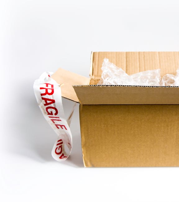 A cardboard box partially open on a plain white surface, with bubble wrap and packing paper visible inside, ready for packing fragile items during a house move. A strip of white packing tape with red 'FRAGILE' lettering is draped over the side of the box. The background shows a clean, neutral environment, suggesting an indoor setting within a property being prepared for a home relocation. The image highlights the careful packing process involved in furniture transport and the importance of proper packaging for fragile antiques, consistent with services offered by Man With a Van Tolworth as part of professional removals and moving logistics.