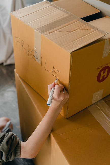 Close-up of a person's hands wrapping an antique item with a smooth, matte green surface in brown packing paper, preparing it for a home relocation. The individual is carefully securing the item inside a cardboard box using crumpled paper for padding, with additional packing materials visible nearby. The scene takes place indoors, with parts of furniture and soft lighting suggesting a residential setting. This detailed packing process, involving protective wrapping and precise handling, is typical of specialist packing options offered by Man With a Van Tolworth for fragile antiques. The careful packaging is part of the overall furniture transport and moving logistics involved in house removals, highlighting the attention to detail necessary for delicate items during a comprehensive moving and packing service.