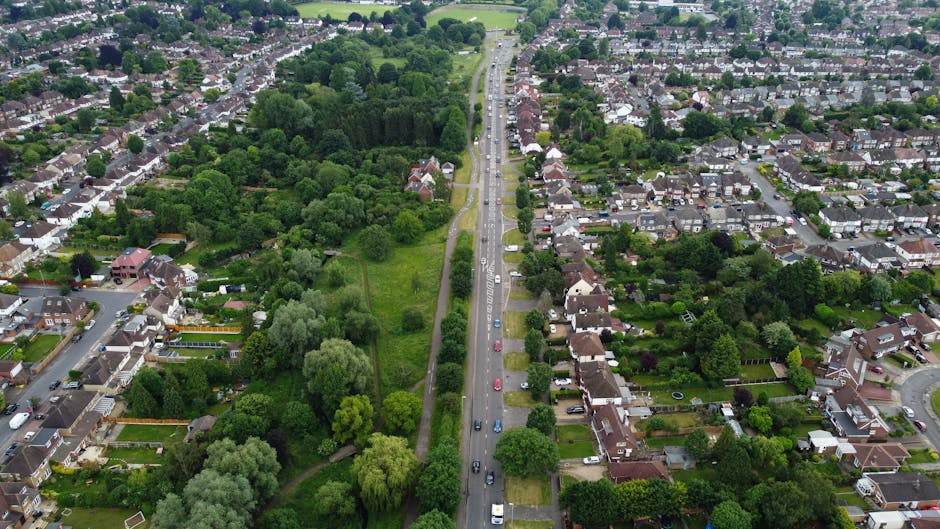 An aerial view of a residential neighbourhood showing a busy main road running through the centre, with cars travelling in both directions and parked along the roadside. To the left of the road, there is a large green park area with dense trees, shrubs, and open grassy spaces. On the right side, rows of terraced and semi-detached houses with gardens, driveways, and various outdoor furniture are visible. Some houses have white or brick exteriors, with roofs covered in slate or tiles, and many gardens are lush with mature trees, bushes, and flowerbeds. The scene is well-lit with natural daylight, indicating a clear day, and the overall setting depicts a peaceful suburban environment suitable for home relocations and moving services offered by Man With a Van Tolworth. The image effectively captures the typical layout of residential and green spaces within the Tolworth area, supporting content related to house removals and moving logistics along the Hook Rise South to Tolworth Court route.