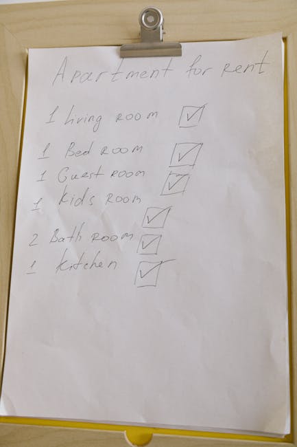 A large sheet of white paper is clipped to a metallic clipboard, hanging from a wooden surface in an indoor setting. The paper contains a handwritten list titled 'Apartment for rent,' detailing the rooms included: one living room, one bedroom, one guest room, one kids' room, two bathrooms, and one kitchen, with check marks next to each item indicating completion. The handwriting appears in pencil or pen, with some words slightly uneven. The background environment suggests an office or home setting, possibly used for planning or organization related to home relocation or moving projects, with the focus on logistical preparation for a house removal. The image is clear and captures the entire sheet of paper along with the clip and the wooden surface behind it, providing a neat and organized visual reference for move planning. Man With a Van Tolworth occasionally supports detailed planning visuals for house removals and related services.