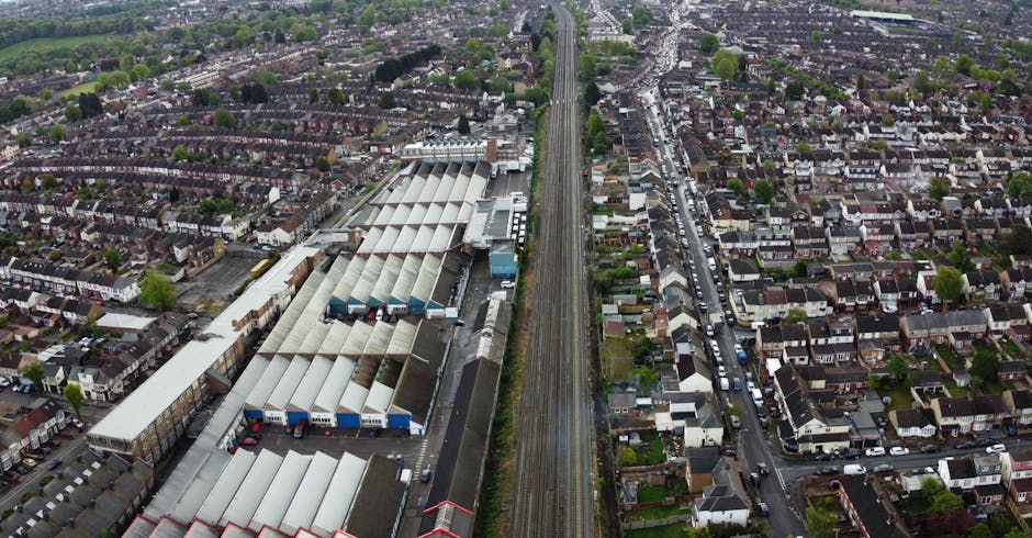 An aerial view of a residential neighbourhood showing a busy main road running through the centre, with cars travelling in both directions and parked along the roadside. To the left of the road, there is a large green park area with dense trees, shrubs, and open grassy spaces. On the right side, rows of terraced and semi-detached houses with gardens, driveways, and various outdoor furniture are visible. Some houses have white or brick exteriors, with roofs covered in slate or tiles, and many gardens are lush with mature trees, bushes, and flowerbeds. The scene is well-lit with natural daylight, indicating a clear day, and the overall setting depicts a peaceful suburban environment suitable for home relocations and moving services offered by Man With a Van Tolworth. The image effectively captures the typical layout of residential and green spaces within the Tolworth area, supporting content related to house removals and moving logistics along the Hook Rise South to Tolworth Court route.