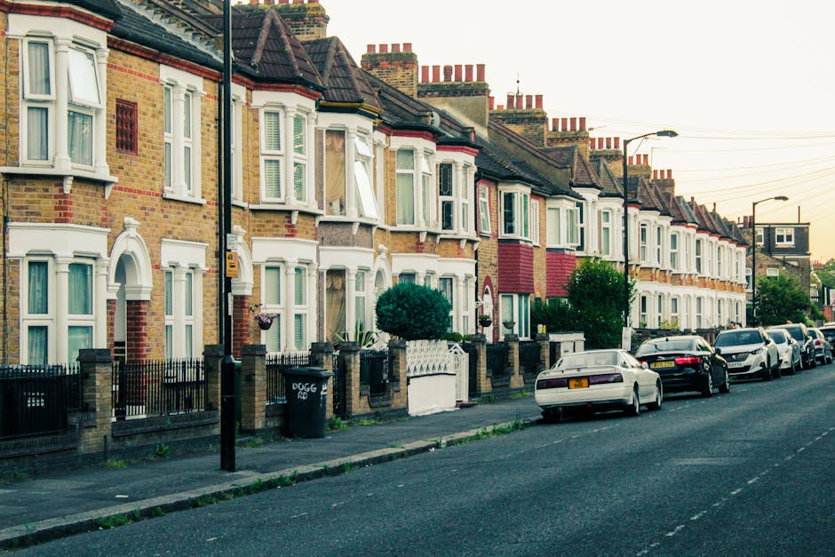 A row of traditional terraced houses with brick facades and bay windows, some open with curtains visible inside, situated along a residential street during daylight hours. In front of the houses, a sidewalk is lined with small gardens enclosed by low brick walls and black metal fences. Parked along the curb are several cars, including sedans and hatchbacks, with one vehicle partially loaded with furniture covered in plastic wrapping and moving blankets, indicating a home relocation process. A moving trolley and packing materials such as cardboard boxes are visible near the vehicle, suggesting ongoing packing or loading activities. Street lamps and overhead power lines extend along the street, and the scene captures a typical suburban residential environment associated with house removals and furniture transport, coordinated by Man With a Van Tolworth during a scheduled move. The lighting suggests late afternoon or early evening, highlighting the quiet, orderly nature of the area.