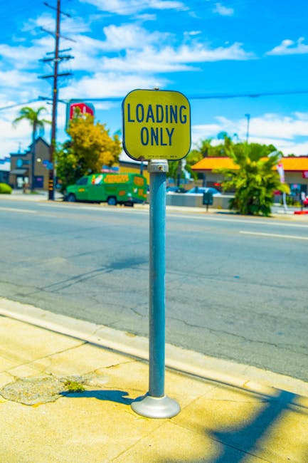 A vertical yellow and black 'Loading Only' parking sign mounted on a metal pole located on a paved sidewalk beside a residential street. In the background, there are power lines, a utility pole, and various buildings including a shop with yellow roof and a green van parked nearby. The sky is blue with scattered white clouds, and nearby trees and greenery are visible. The sign indicates designated loading and unloading areas, which can be relevant for home relocation or furniture transport during moving services. This image illustrates the type of parking restriction that logistical planning by [COMPANY_NAME] such as Man With a Van Tolworth would consider when arranging efficient loading and unloading points for house removals along Ewell Road, TOLWORTH.