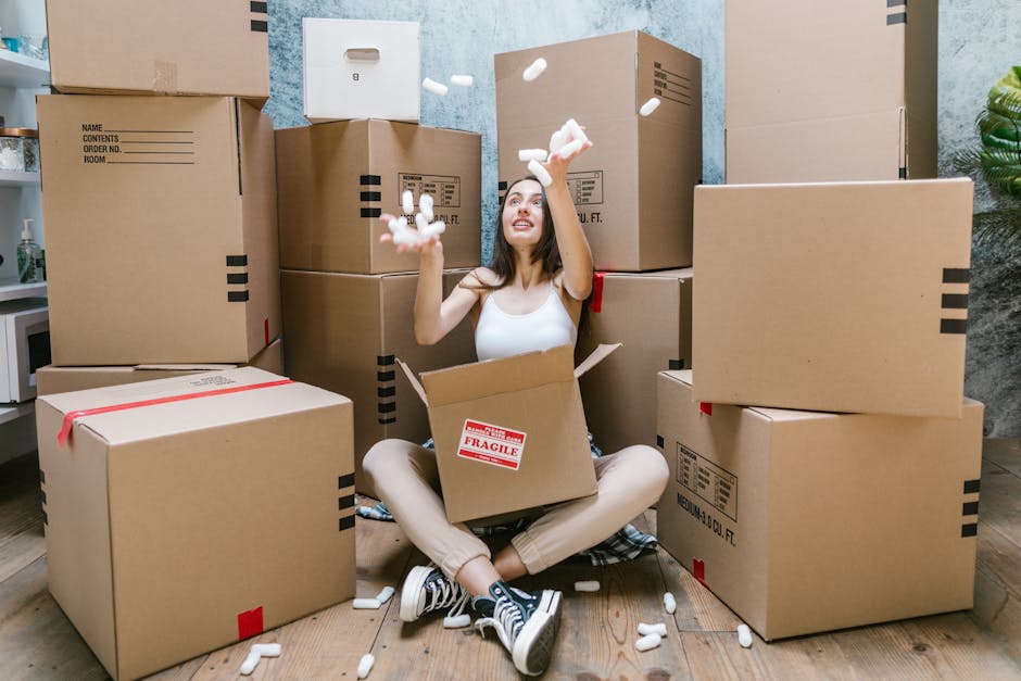 A young woman sitting cross-legged on a wooden floor inside a room, surrounded by large cardboard moving boxes in various sizes, some sealed with red tape and labeled with packing information. She is wearing casual clothing with striped sneakers and is smiling while tossing packing foam peanuts into the air, indicating a packing or moving process. Behind her, there is a light-colored wall and a small potted plant, with additional boxes stacked against the wall. The scene captures the packing stage of a home relocation, with the boxes organized for transport, as part of a furniture transport or moving service provided by Man With a Van Tolworth, supporting house removals and logistics for the street-by-street removals guide in Tolworth.