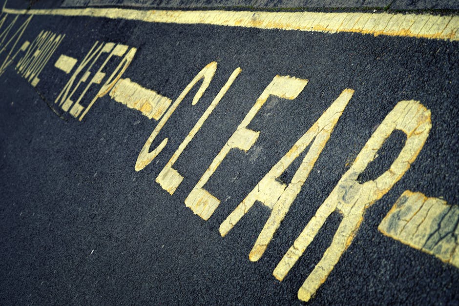 Close-up photograph of a weathered yellow painted 'KEEP CLEAR' warning on a dark asphalt pavement, with visible cracks and peeling paint, situated outside a property entrance used for home removals. The image shows a section of the roadway with the painted message oriented diagonally, emphasizing its importance in parking and access regulations during the furniture transport or loading process. In the background, a partially visible curb and a roadside surface can be seen, indicating an outdoor setting near residential or commercial premises. This scene is representative of typical parking restriction markings encountered during house relocation tasks in Tolworth, where clear access for removals vehicles is essential. Man With a Van Tolworth, operating within the removals service category, ensures adherence to parking and pavement rules when managing home moves and furniture transport, supporting smooth logistical operations on moving day.
