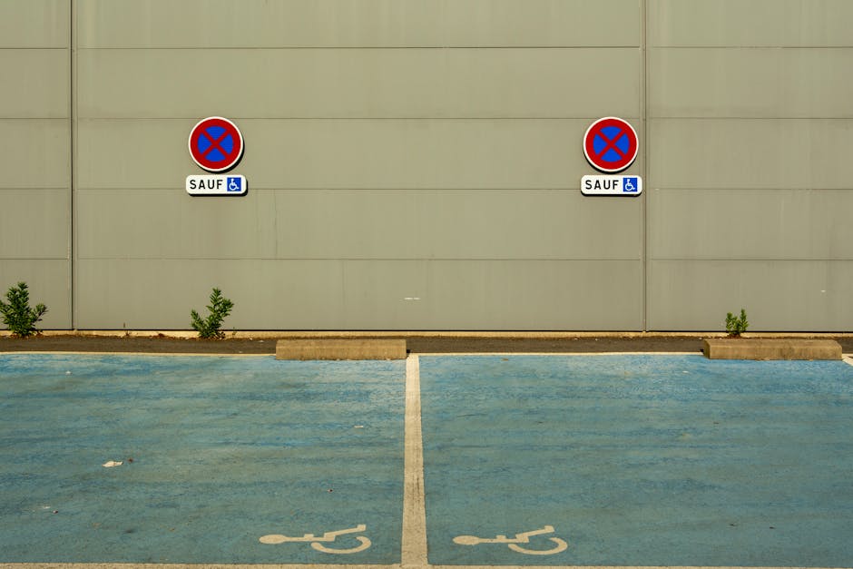 An empty parking space designated for disabled individuals, marked with blue painted wheelchair symbols on the asphalt. The parking area is situated in front of a beige building wall, which features two no-parking signs with red circles and blue backgrounds, each accompanied by a small blue sign indicating accessibility. The signs are mounted on the wall at a uniform height and are positioned above the parking space, which is separated by concrete wheel stops. Small green plants grow between the pavement and the building wall, adding slight greenery to the scene. The overall environment is well-lit, indicating daytime, and the space appears clean and ready for vehicle loading or home relocation activities, relevant to the logistics of removals and furniture transport managed by [COMPANY_NAME].