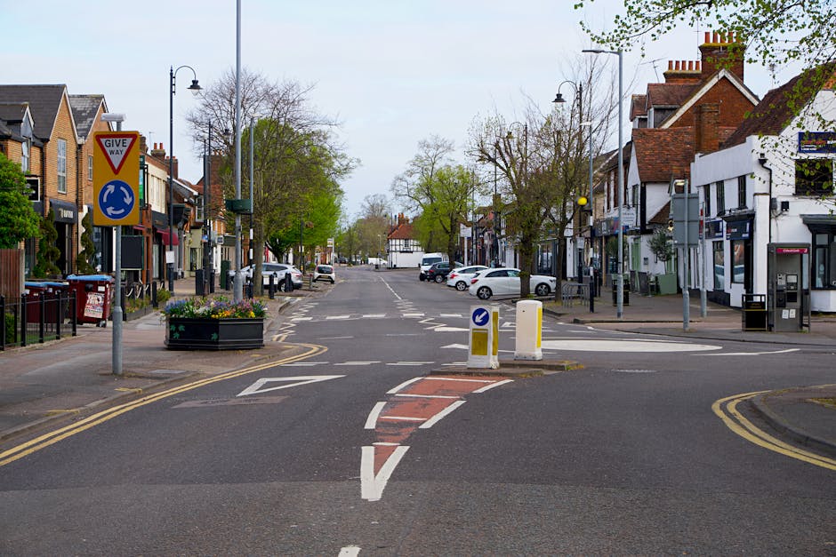 A quiet urban street scene showing a T-junction with white dashed and solid lines, a give way sign, and a roundabout sign. The intersection is surrounded by a mix of residential and commercial buildin