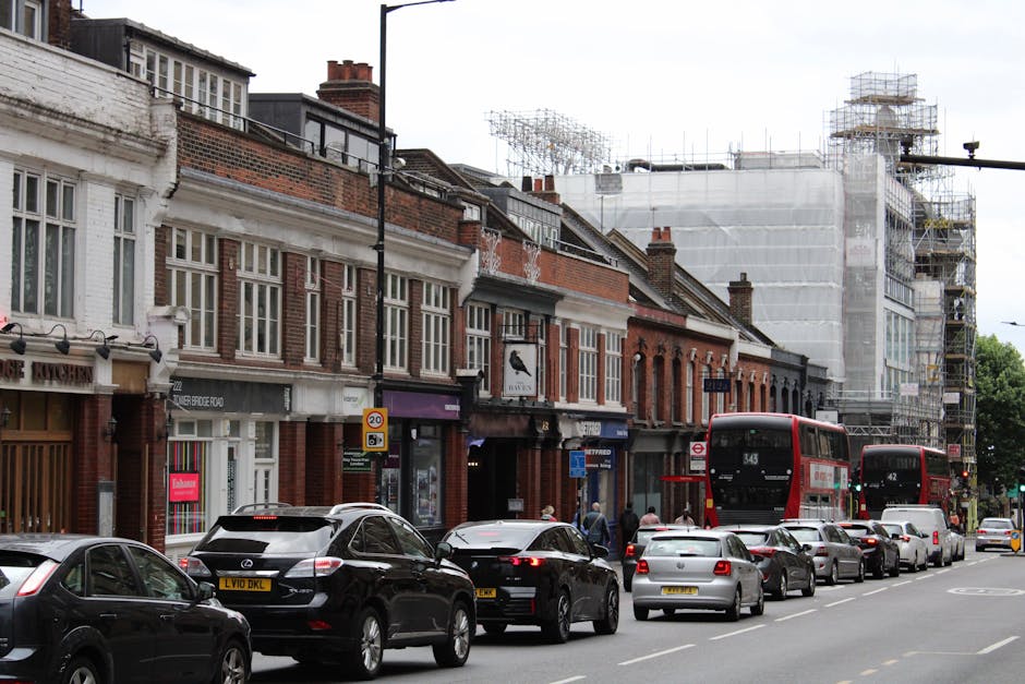 A quiet urban street scene showing a T-junction with white dashed and solid lines, a give way sign, and a roundabout sign. The intersection is surrounded by a mix of residential and commercial buildin