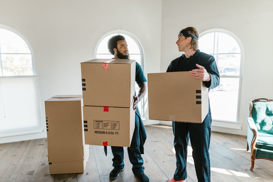 Two men are inside a bright room preparing for a house move, with large arched windows allowing natural light to illuminate the space. One man, with a beard and curly hair, is holding a medium-sized cardboard box with red tape, while the other man, wearing a headband, is carrying a smaller cardboard box. Several other packing cartons, some sealed with tape and labeled, are stacked on the wooden floor nearby. The room features minimal furniture, including a vintage-style green upholstered armchair with wooden arms and legs positioned against the wall near the windows. The men are dressed in casual, practical clothing suitable for furniture transport and packing, with one wearing dark trousers and the other in black pants. The scene captures the logistics involved in packing and loading household items for a professional removals service, such as those offered by Man With a Van Tolworth, preparing for house relocation or moving day, with equipment like boxes and possibly moving blankets or trolleys out of view.
