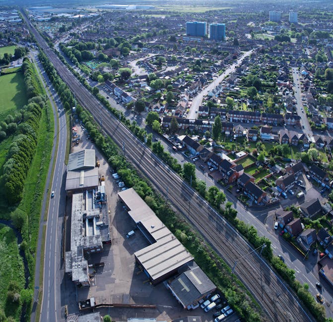 An aerial view of a suburban area showing a railway line running parallel to a road, with residential houses on one side and a large industrial or warehouse building in the foreground. The scene includes parked vehicles, trees lining the roads, and distant high-rise buildings on the horizon. The image captures the urban landscape and transportation infrastructure, relevant to house removals and moving logistics, with natural lighting highlighting the structural details of the buildings and greenery. This view exemplifies the environment where Man With a Van Tolworth provides home relocation and furniture transport services, including loading and packing operations near the railway and roadways.