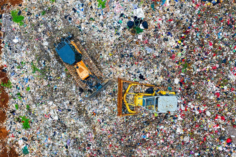 An aerial view of a large landfill site filled with mixed waste, including plastic bottles, cardboard, textiles, and other household rubbish. Two tracked excavators are visible; one yellow and one orange, actively engaged in moving and spreading the waste materials across the site. The excavators are positioned on uneven terrain covered with garbage, with their buckets scooping and shifting debris. Near the top right, there are scattered items such as black plastic bins, fabric or clothing pieces, and assorted rubbish. The scene is outdoors, with natural lighting illuminating the dense collection of waste, emphasizing the scale and mess of the disposal area. This image illustrates the process of waste management and disposal, relevant to home relocation and wrapping or packing waste, as managed by companies like Man With a Van Tolworth, especially for complying with waste disposal laws after moving in Tolworth (KT6).