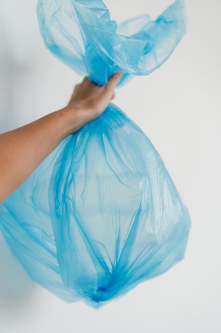 A person's hand holding a blue plastic waste disposal bag, which is partially filled and twisted at the top. The bag appears to contain domestic rubbish, including paper and plastic waste. The background is plain and white, with soft lighting illuminating the scene. This image demonstrates proper waste disposal in the context of house removals or home relocation. It is relevant to services such as packing and moving, where efficient disposal of waste is part of the process. Man With a Van Tolworth offers removals that include responsible waste management during relocation, ensuring compliance with local waste disposal laws after moving in Tolworth (KT6). The focus on waste collection and disposal is integral to the logistics of a smooth moving process, highlighting the importance of proper debris handling during home or office removals.